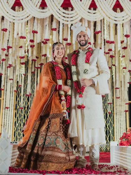 A happy portrait of the couple post-ceremony. The rich, traditional colors of their attire contrast beautifully with the ivory and red decor of the mandap.