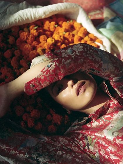 A close-up portrait from the 'Phool Mandi' editorial. The model lies amidst a pile of marigolds, with the rich red and gold of her patterned outfit creating a beautiful texture and color harmony with the flowers.