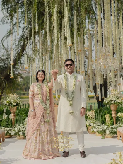 The couple raises their hands in a gesture of celebration after their Jaimala. The backdrop of hanging white florals from the trees creates a stunning, ethereal effect.