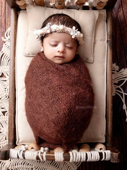 A top-down view of a baby in a brown swaddle and floral crown, sleeping on a miniature wooden bed. The contrast of dark wood and soft textures is beautiful.