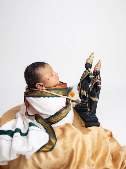 A color version of the devotional pose, with the baby wrapped in a traditional white dhoti with a green and gold border, looking towards the idols.
