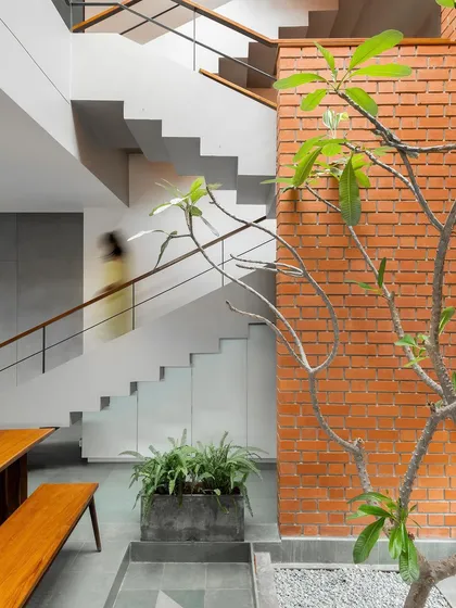 The main staircase is a prominent feature, with its clean white lines contrasting against the textured brick of the courtyard wall. A frangipani tree grows in the courtyard, its branches reaching up towards the skylight.