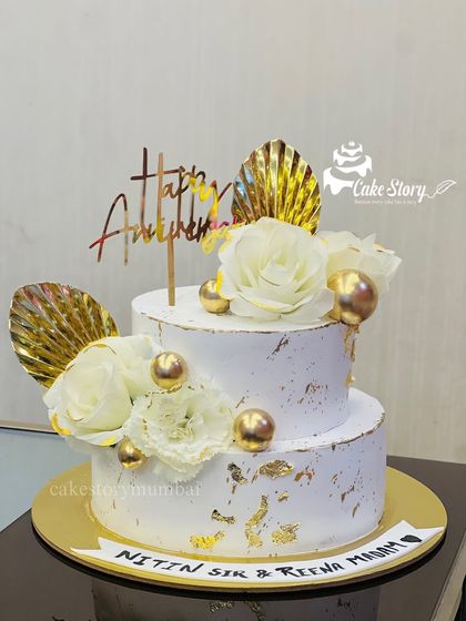 A stunning white and gold anniversary cake. This two-tier design is decorated with white roses, gold palm leaves, and gold leaf flakes for a touch of modern elegance.