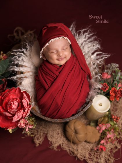 A baby smiling in their sleep, swaddled in a rich red wrap and sleepy hat. The rustic, warm-toned props create a cozy and inviting atmosphere.