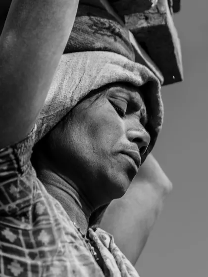 A close-up black and white portrait of a woman carrying bricks, her eyes closed in a moment of exertion and concentration.