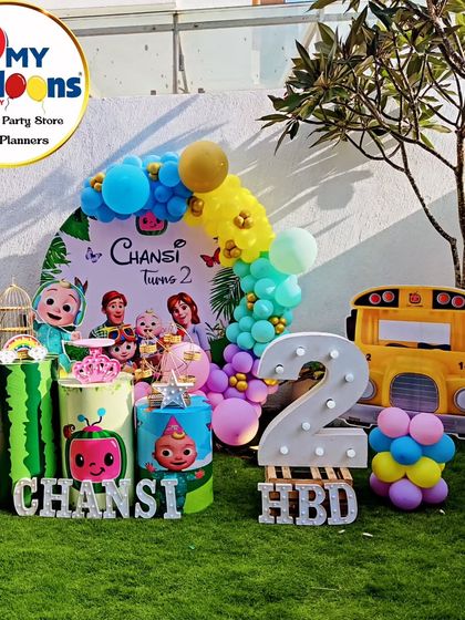 An outdoor Cocomelon-themed second birthday party. The setup includes a character backdrop, themed plinths, and a large light-up number '2' set on a lush green lawn.