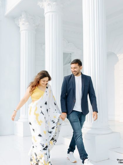A happy, carefree moment during a pre-wedding shoot. The couple walks hand-in-hand through a beautiful white colonnade, their smiles lighting up the frame.