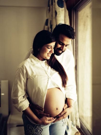 A warm and loving indoor portrait. The couple embraces by a window, the natural light illuminating them as they both cradle the baby bump.