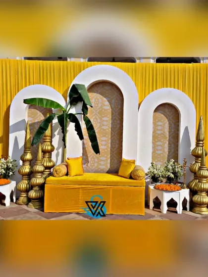 The main seating area for a Haldi function, featuring a custom-made yellow sofa set against a backdrop of modern arches and traditional brass elements. The banana leaves add a touch of tropical tradition.