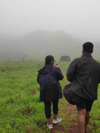 Two trekkers walking towards the misty hills of Kodachadri, with ancient structures dotting the landscape.