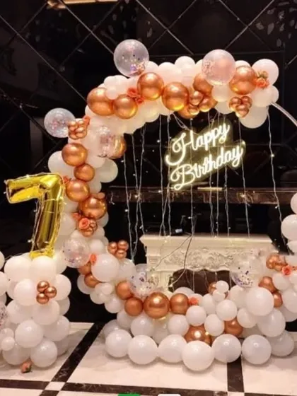 A 67th birthday party with a circular balloon arch in white and rose gold. The setup includes fairy lights, a neon sign, and foil number balloons.