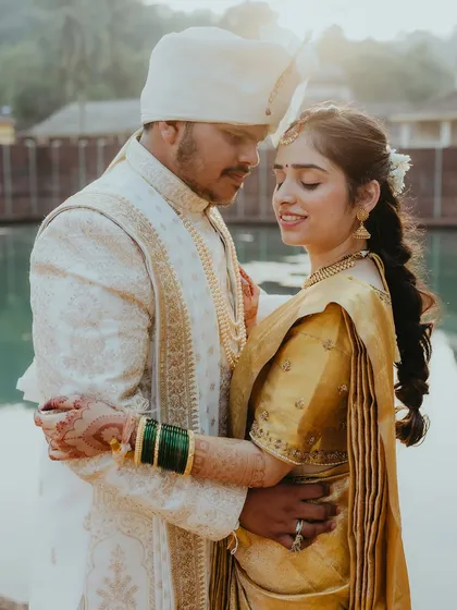 A beautiful, sun-kissed portrait of a Konkani couple in Goa, sharing a quiet moment by the temple pond. The warm light highlights the richness of their traditional wedding attire.