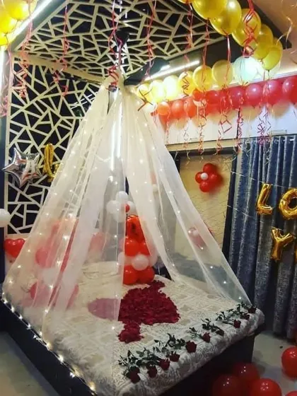 An intimate room setup featuring a canopy tent over the bed, decorated with fairy lights and rose petals. 'I LOVE YOU' foil letters add to the romantic message.