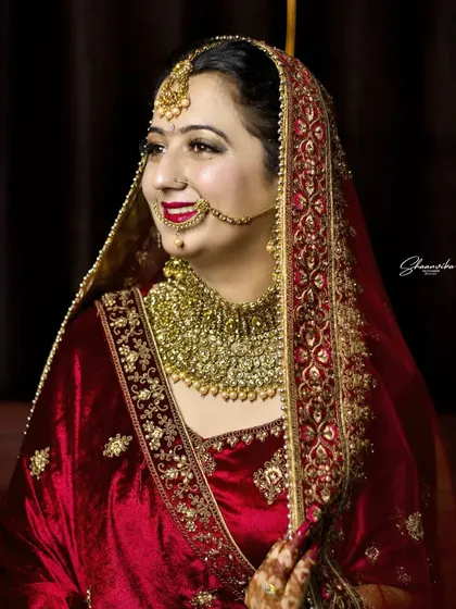 A candid moment of a bride smiling, captured in her beautiful red velvet wedding outfit. The heavy gold jewelry and soft lighting create a classic, regal bridal portrait.