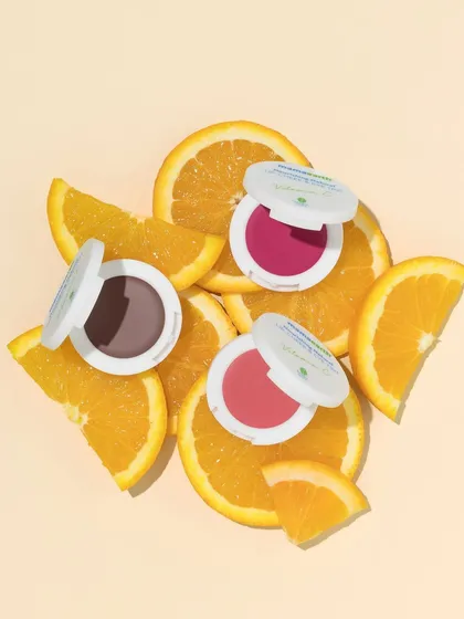 A vibrant flatlay featuring lip and cheek tints nestled among fresh orange slices. The composition is bright and energetic, perfectly communicating the product's Vitamin C infusion and fruity vibe.