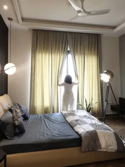 This warm and cozy bedroom is designed to welcome the morning sun, with large windows and light curtains. The room features a dark wood fluted panel wall behind the bed, which contrasts beautifully with the soft, neutral tones of the furnishings.