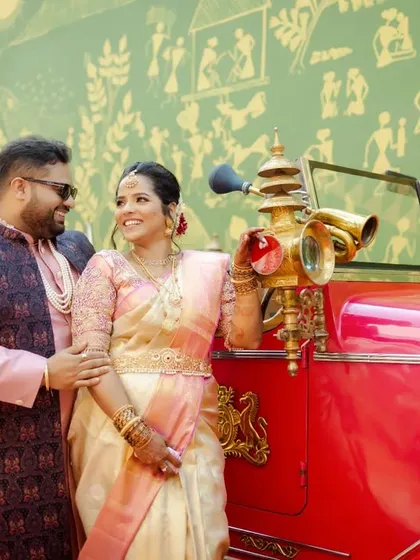 A classic portrait of the couple with their vintage red wedding car, set against a traditional Warli painting wall.