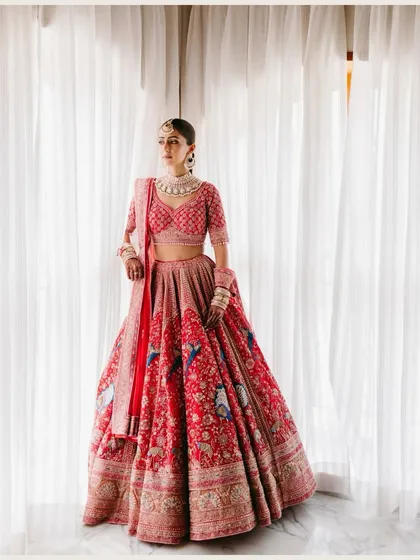 A full-length portrait of a bride in a magnificent red lehenga, standing against a white curtain that diffuses the light beautifully, creating a classic and regal look.