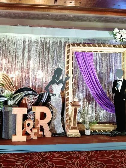 A glamorous photo booth designed for a sangeet. It features a shimmering silver backdrop, a large golden frame, and stylish cutouts of a couple, perfect for fun pictures.