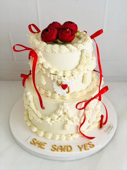A vintage-inspired "She Said Yes" cake. This two-tier design uses intricate piping and delicate red ribbons to create a timeless and romantic look for an engagement celebration.