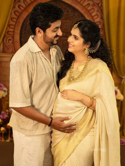 A joyful couple's portrait with traditional styling. The expecting mother, in a cream and gold saree, and her partner share a happy look in a royally decorated setting.