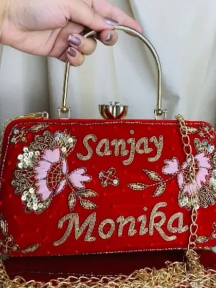 This red velvet clutch features delicate pink and gold floral embroidery alongside the couple's names, 'Sanjay' and 'Monika'. It also comes with a detachable gold chain strap for convenience.