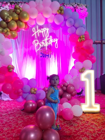 A happy moment from a first birthday party we decorated. The setup includes a custom neon 'Happy Birthday' sign, a balloon arch in pink and gold, and our popular light-up '1' marquee number prop.