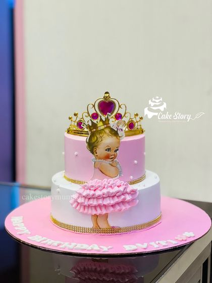 A royal cake for a little princess. This two-tier design features a baby princess photo topper, a golden crown, and a delicate pink tutu made of buttercream ruffles.