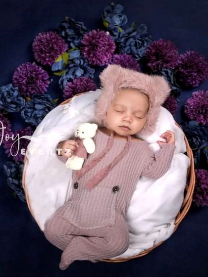 A cozy teddy bear theme featuring a newborn in a buttoned romper and a furry bear hat. The baby holds a tiny teddy bear while resting in a basket, surrounded by deep purple flowers.