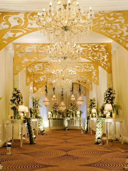 A grand, golden-themed entrance corridor for a luxurious reception. The design features intricate gold laser-cut panels, tall floral arrangements, and warm lighting to create an opulent atmosphere.
