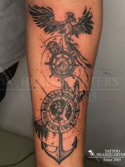 A large forearm piece combining a phoenix, a ship's wheel, a compass, and an anchor, telling a story of travel, guidance, and rebirth.