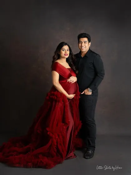 A classic and elegant couple's portrait. The mom-to-be is dressed in a stunning dark red ruffled gown, posing with her partner against a simple, dark studio background.