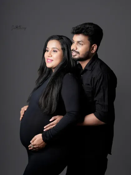 A beautiful and classic couple portrait in matching black outfits. The husband stands behind his wife, gently holding her bump, creating a timeless image of support and love.