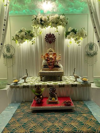 An ethereal all-white Ganpati decoration. The setup includes floating cloud-like flower arrangements, dreamcatchers, and soft green lighting to create a peaceful and divine space.