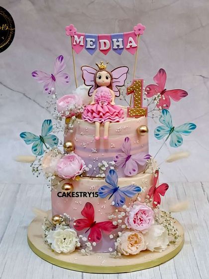 A dreamy two-tier red velvet cake in soft lavender hues for a first birthday. This doll-themed creation is adorned with delicate butterflies, fresh flowers, and a custom name banner for a personal touch.