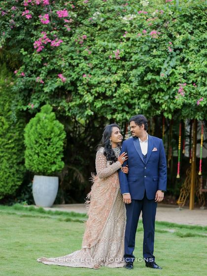 A full-length shot of a couple in a green garden, showcasing her beautiful traditional lehenga and his coordinating suit.