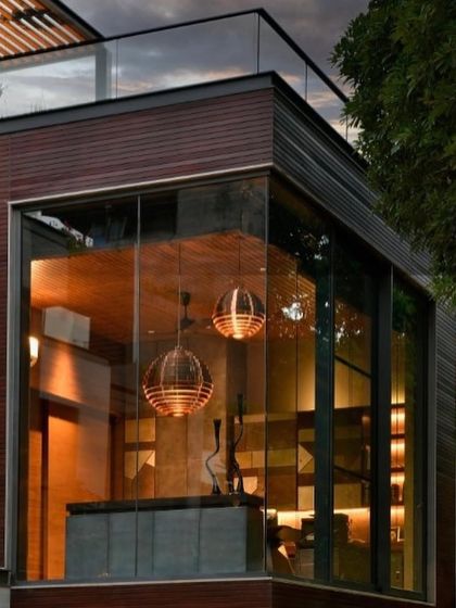 An exterior view of the cantilevered study at T House. The glass corner box appears to float among the trees, and the warm glow from the copper pendant lights within creates a jewel-like effect at dusk.