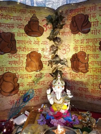 This artistic Ganpati setup uses a hand-painted backdrop resembling a sacred text, with depictions of the eight forms of Ganesha. The idol is placed at the center, surrounded by offerings and a peacock feather.