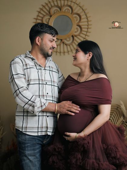 A sweet moment between the couple during their maternity session. The partner gazes lovingly at the mother-to-be, who is dressed in an elegant maroon gown, his hand resting on her bump.