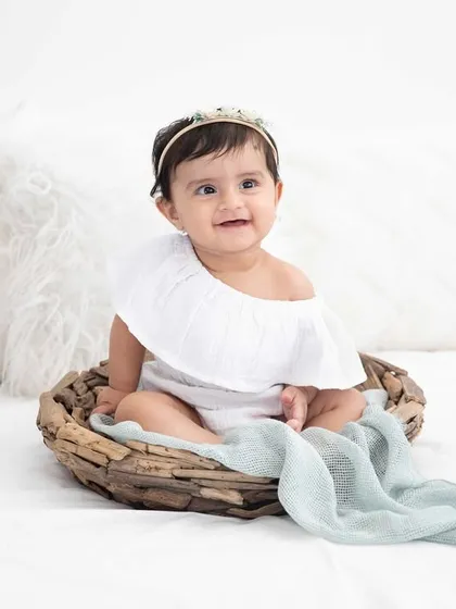 A happy baby girl with a sweet headband, sitting in a rustic basket on a bed. Her bright smile lights up this simple and classic portrait.