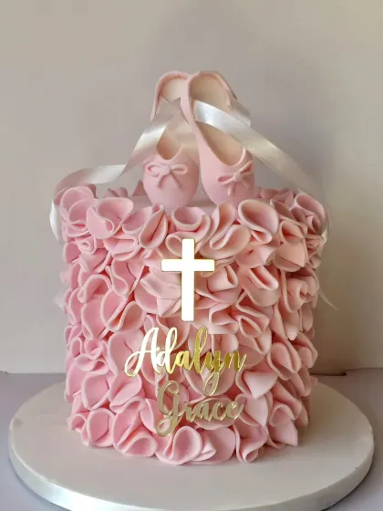This christening cake is all about delicate textures and symbols. The pink ruffled fondant creates a soft, fabric-like effect, adorned with a simple cross and the baby's name. The edible baby ballet shoes on top add a touch of sweetness.
