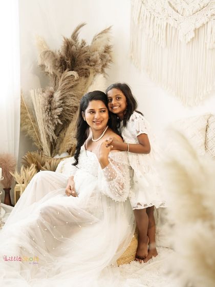 A mother and daughter moment in a dreamy, light-filled boho setting. The daughter stands behind her seated mother, both in white, sharing a sweet and gentle connection.