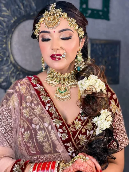 A beautiful bridal portrait with a focus on the complete look. The bride wears a maroon lehenga with heavy jewelry, and her makeup features a bold red lip and shimmering eyes, perfect for her wedding day.