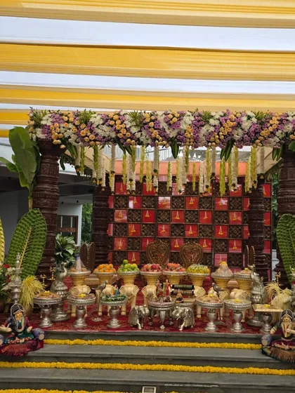A grand outdoor mandapam with a colorful yellow and white canopy. The stage features Chettinad-style pillars, a traditional floral backdrop, and large banana leaf decorations.