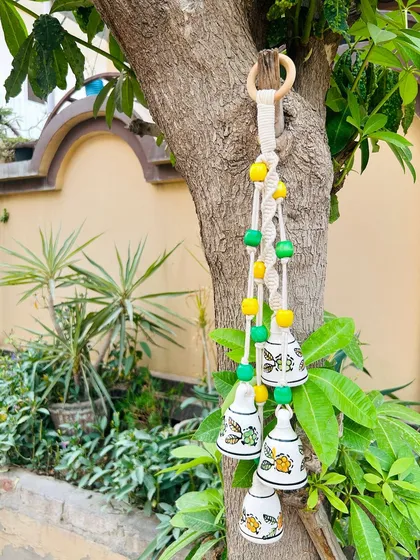 A close-up of our 'Symphony Chimes' in the 'Amaltas' variant, hanging from a tree. The combination of macrame knots, colorful wooden beads, and ceramic bells makes for a beautiful outdoor decor piece.