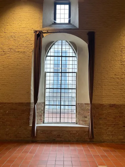 A magnificent arched window inside the Belfort Museum in Bruges. The scale of the window, combined with the exposed brick wall and terracotta floor, creates a powerful sense of history and space.