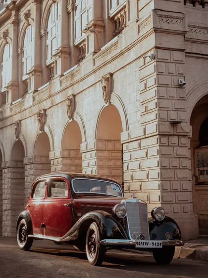 A classic two-tone burgundy and black Mercedes-Benz sedan from a bygone era, parked on a city street. The warm evening light highlights its vintage design against the grand, stone architecture of a heritage building in Mumbai.