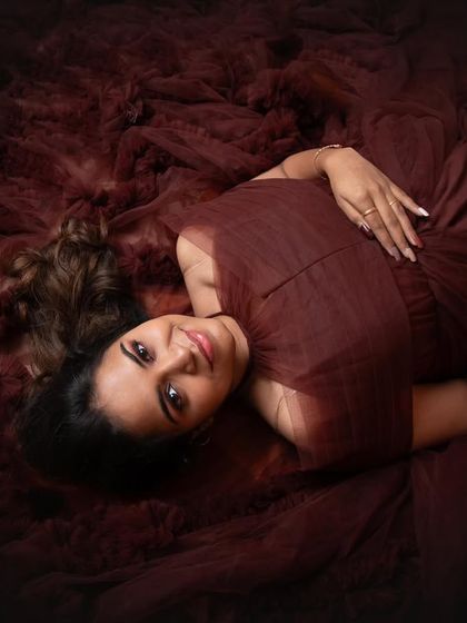 A gorgeous overhead shot. The mom-to-be lies on the rich, textured fabric of her gown, creating a dramatic and artistic "sleeping beauty" style portrait.