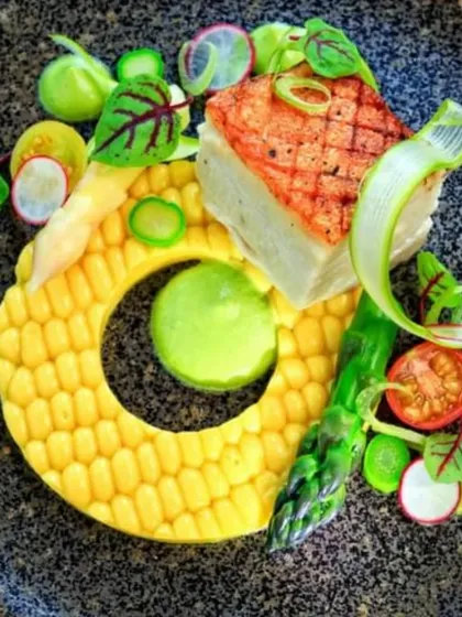 We love playing with textures and shapes in our plating. This dish features a grilled piece of fish served with an artistic corn-kernel ring, asparagus, and fresh greens, creating a visually stunning plate.