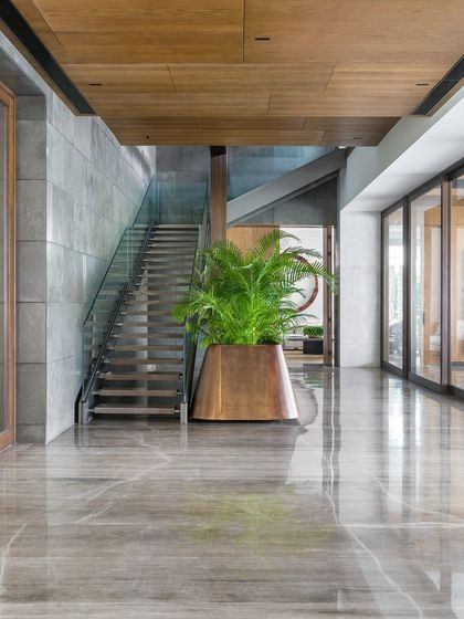 A view down the expansive lobby towards the metal and marble staircase. A large, custom brass planter adds a touch of nature, drawing the eye forward and highlighting the home's impressive scale and double-height areas.